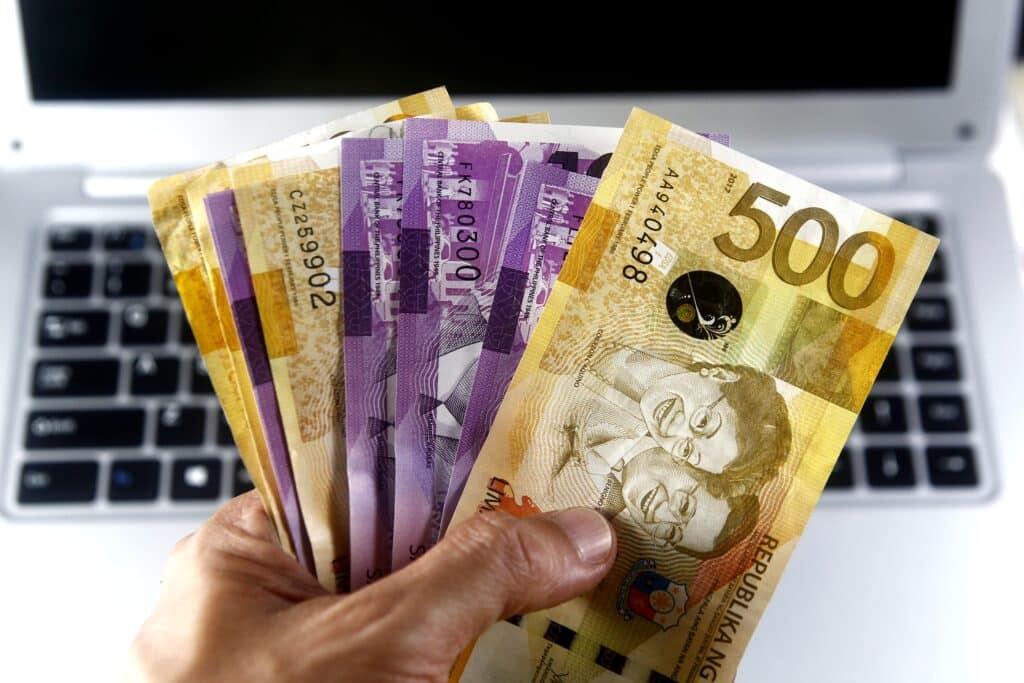 A closeup of a hand holding Philippine currency in front of a laptop