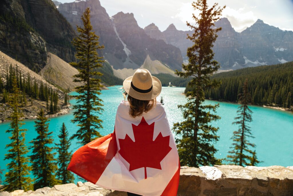 Une femme enveloppée par un drapeau canadien qui admire le paysage d'un lac et des montagnes allentours