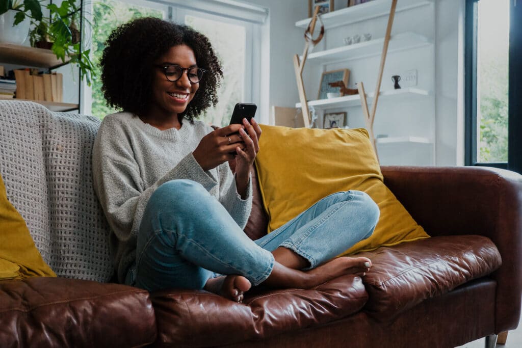 Currencies of the world: woman happily using her phone while sitting on her couch
