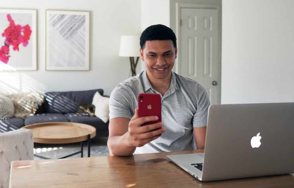 Une homme devant son ordinateur portable souriant en utilisant son téléphone portable