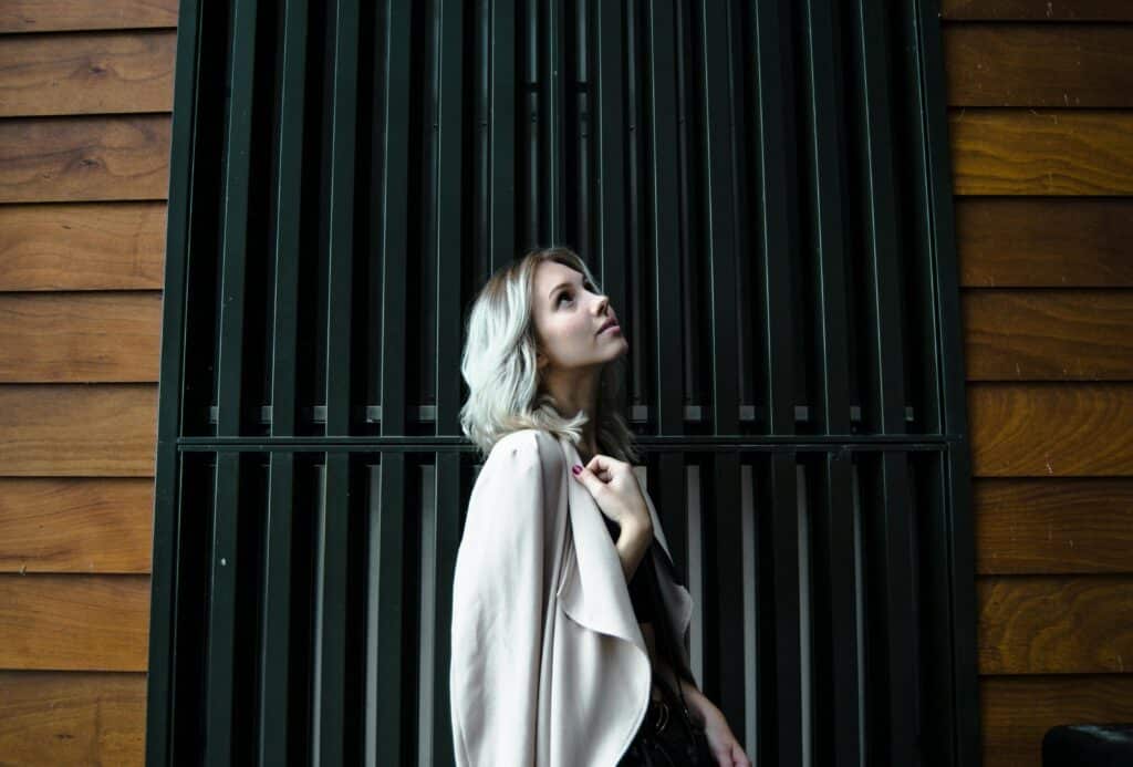 Side View Photo of Woman in White Coat Standing In Front of Black Metal Door Looking Up in Tallinn, Harju County, Estonia